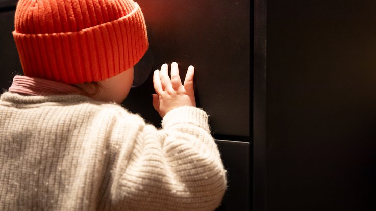 VISUELL Scenography: Permanent exhibition at Hornmoldhaus: Detail of a child wearing a red cap looking through an integrated peephole in a black display cabinet. The child's hand is resting on the surface of the interactive station.