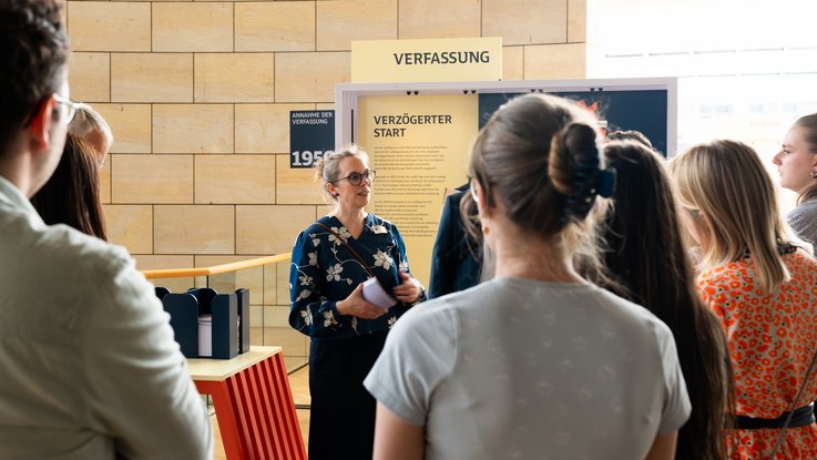 VISUELL Szenografie: Wanderausstellung „Jubiläum. 75 Jahre Verfassung NRW“ für die Stiftung Haus der Geschichte Nordrhein-Westfalen VISUELL Szenografie: Wanderausstellung: Gruppenführung vor dem Ausstellungselement „Verfassung – Verzögerter Start“. Eine Frau erklärt Inhalte vor gelber Wand mit Jahreszahl 1950.