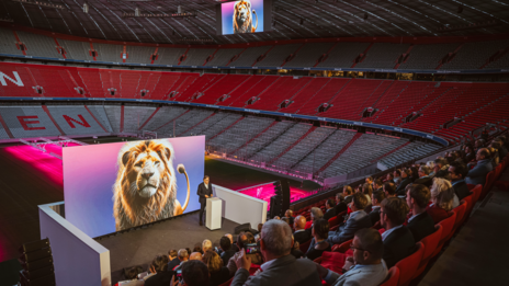 VISUELL Szenografie: Event „NEXT LEVEL“: Die Zuschauenden sitzen auf der Tribüne, eine Person steht hinter einem Rednerpult vor der großen LED-Leinwand. Im Hintergrund sieht man die leeren ränge der Allianz Arena und das Spielfeld.