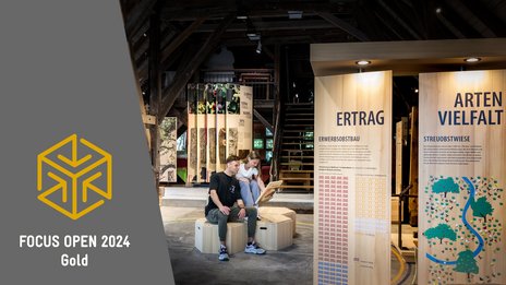 VISUELL Szenografie: Obstbaumuseum: Zwei Besucher sitzen in der Mitte der Ausstellung und betrachten eine Informationstafel in ihren Händen