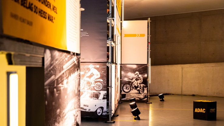 VISUELL Szenografie: Ausstellung: Perspektive durch die Ausstellungsarchitektur; schwarz-weiße Motorsportfotos auf Bannern treffen auf klare gelbe Flächen und gezielte Lichtinszenierung.