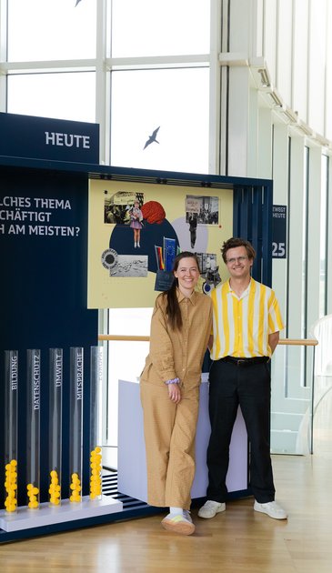 VISUELL Szenografie: Wanderausstellung „Jubiläum. 75 Jahre Verfassung NRW“ für die Stiftung Haus der Geschichte Nordrhein-Westfalen VISUELL Szenografie: Wanderausstellung: Die Geschäftsführerin und der Geschäftsführer stehen lächelnd vor dem Modul „Heute“. Hinter ihnen Illustrationen und Themenfragen zur Gegenwart, davor transparente Röhren mit gelben Kugeln zur Abstimmung über Themenbereiche.