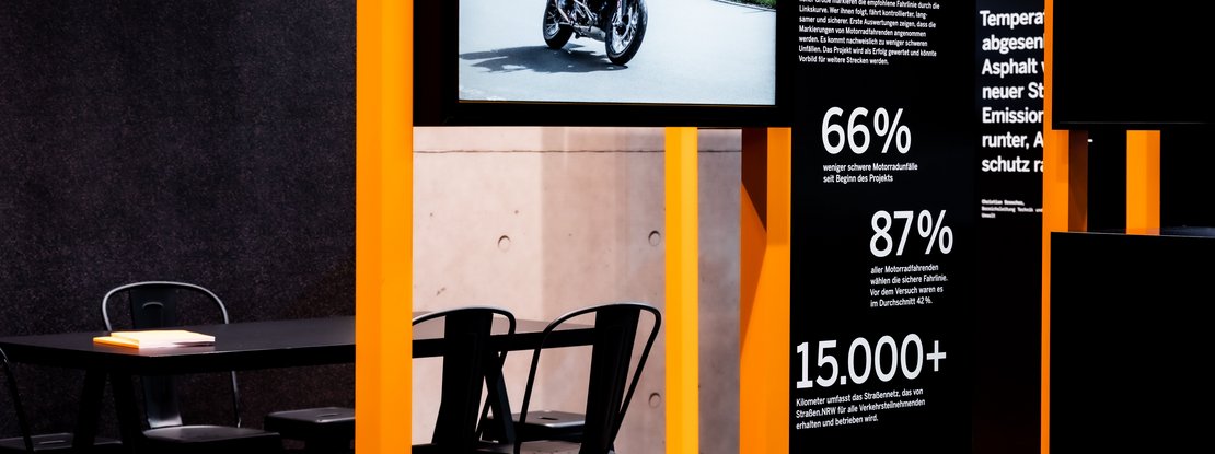 VISUELL Scenography: Straßen.NRW stand at InfraTech 2026: The image shows a modular exhibition system and a table with chairs behind it; an integrated screen displays a thematic film, flanked by information panels featuring striking typography and statistical data.