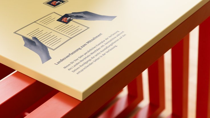 VISUELL Szenografie: Wanderausstellung „Jubiläum. 75 Jahre Verfassung NRW“ für die Stiftung Haus der Geschichte Nordrhein-Westfalen VISUELL Szenografie: Wanderausstellung: Detail einer gelben Tischfläche mit grafischer Illustration einer aufgeschlagenen Broschüre und dem Hinweis „Landesverfassung zum Mitnehmen!“.