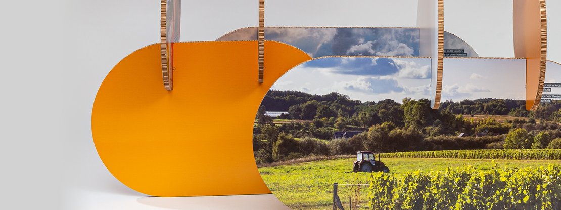 VISUELL Szenografie: Wanderausstellung Robert Bosch Stiftung GmbH: Panoramatische Teilansicht eines modularen Ausstellungselements mit organisch geschwungenen, orangefarbenen Formen und integrierten Landschaftsfotografien; senkrechte und waagerechte Elemente prägen die Komposition.