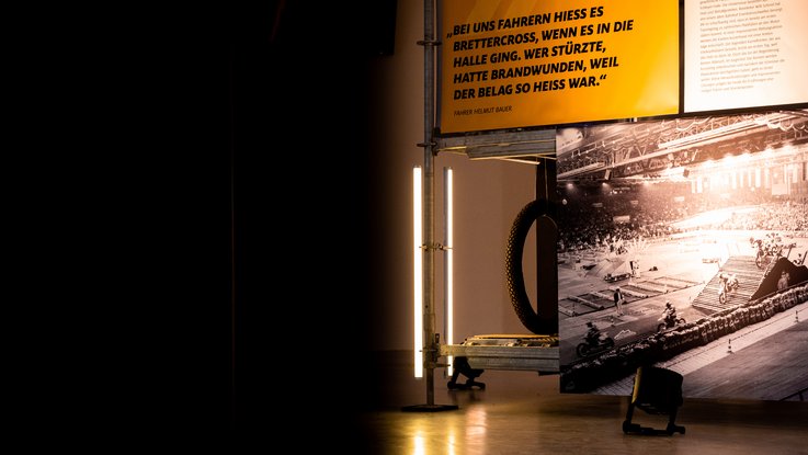 VISUELL Szenografie: Ausstellung: Stimmungsvolle Ansicht mit gelb leuchtender Texttafel und historischem Schwarz-Weiß-Foto einer Indoor-Rennszene; Reifen und Gerüstkonstruktion rahmen das Motiv.