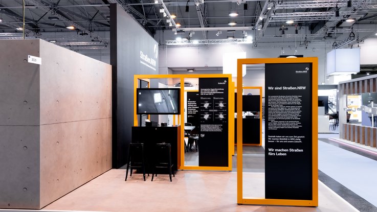 VISUELL Scenography: Straßen.NRW stand at InfraTech 2026: A perspective view of the stand design; in the foreground is an information pillar bearing the headline “We build roads for life”, whilst behind it, modular frames structure the space for in-depth discussions with experts.