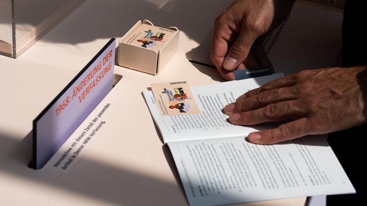 VISUELL Szenografie: Wanderausstellung „Jubiläum. 75 Jahre Verfassung NRW“ für die Stiftung Haus der Geschichte Nordrhein-Westfalen VISUELL Szenografie: Wanderausstellung: Hände einer Person legen eine Themenkarte mit Aufschrift „Bildung“ in ein aufgeschlagenes Verfassungsheft – interaktives Element der Ausstellung.