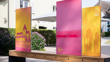 VISUELL Szenografie: Wahre Wunderbänke: Rückseite einer Sitzbank mit Informationstafeln mit drei plakativen Bildmotiven der Klosterinsel Reichenau in magenta, gelb und orange