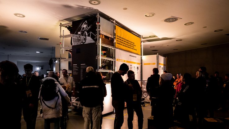 VISUELL Szenografie: Ausstellung: Besucherinnen und Besucher bewegen sich durch die Ausstellungsarchitektur mit Bannern, Fotos und Zitaten; die helle Inszenierung hebt sich vom dunklen Umfeld ab.