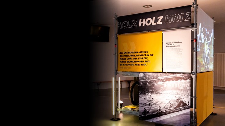 VISUELL Szenografie: Ausstellung: Kubus mit plakativer Aufschrift „HOLZ HOLZ HOLZ“; darunter großformatige Fotos und Zitatbanner, die historische Perspektiven vermitteln.