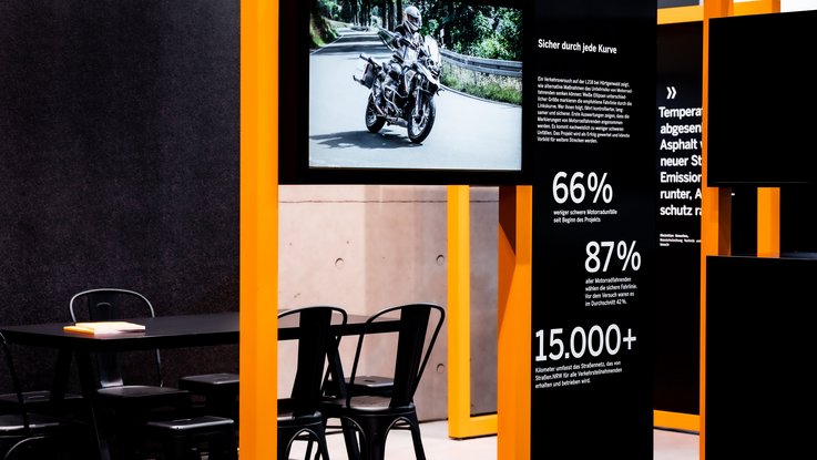 VISUELL Szenografie: Messestand Straßen.NRW InfraTech 2026: Ausschnitt zeigt modulares Ausstellungssystem sowie einen Tisch mit Stühlen dahinter; ein integrierter Screen zeigt einen Themenfilm, flankiert von Infopaneelen mit prägnanter Typografie und statistischen Daten.