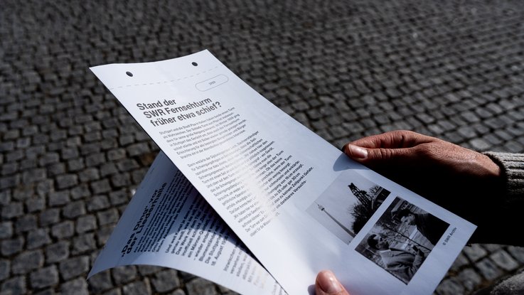 VISUELL Szenografie: Jubiläumsausstellung: 70 Jahre Fernsehturm und Liederhalle: Besucher hält ein Informationsblatt mit historischen Fotografien und architektonischen Hintergründen zu Fernsehturm und Liederhalle in der Hand.