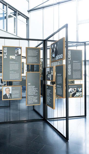VISUELL Szenografie: Jubiläumsausstellung: In einer Rahmenkonstruktion aus Metall sind an Drahtseilen Informationstafeln in Holzrahmen in verschiedenen Formaten befestigt.
