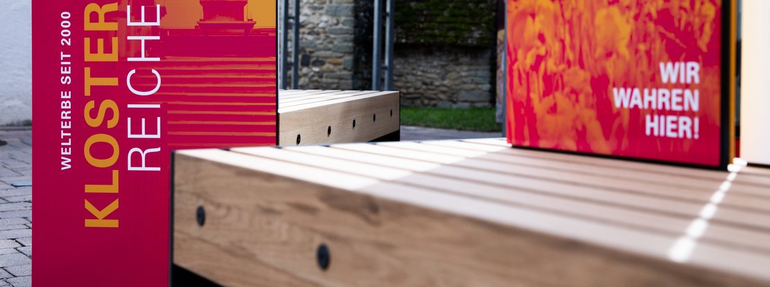 VISUELL Szenografie: Wahre Wunderbänke: Nahaufnahme der Sitzbänke mit Informationseinheiten der Hauptansichtsseite mit plakativen Bildmotiven in magenta und orange mit dem Titel der Welterbestätte sowie der Aufschrift WIR WAHREN HIER!
