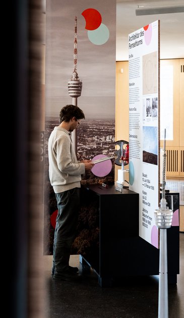 VISUELL Szenografie: Jubiläumsausstellung: 70 Jahre Fernsehturm und Liederhalle: Besucher betrachtet ein interaktives, haptisches Ausstellungselement umgeben von weiteren Informationswänden zur Geschichte der beiden Gebäude.