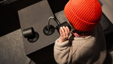 VISUELL Scenography: Permanent exhibition at Hornmoldhaus: Detail of a listening station with two integrated loudspeakers on a black exhibition table. A child wearing a red cap uses the flexibly mounted headphones to listen to the history of the city.
