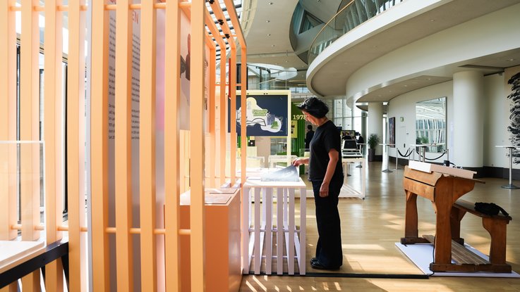 VISUELL Szenografie: Wanderausstellung „Jubiläum. 75 Jahre Verfassung NRW“ für die Stiftung Haus der Geschichte Nordrhein-Westfalen VISUELL Szenografie: Wanderausstellung: Besucherin betrachtet Exponate an einem orangefarbenen Modul, im Hintergrund weitere Themenstationen wie Umwelt und Bildung in der lichtdurchfluteten Ausstellungshalle.