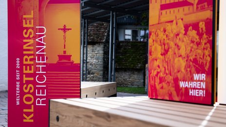 VISUELL Szenografie: Wahre Wunderbänke: Nahaufnahme der Sitzbänke mit Informationseinheiten der Hauptansichtsseite mit plakativen Bildmotiven in magenta und orange mit dem Titel der Welterbestätte sowie der Aufschrift WIR WAHREN HIER!