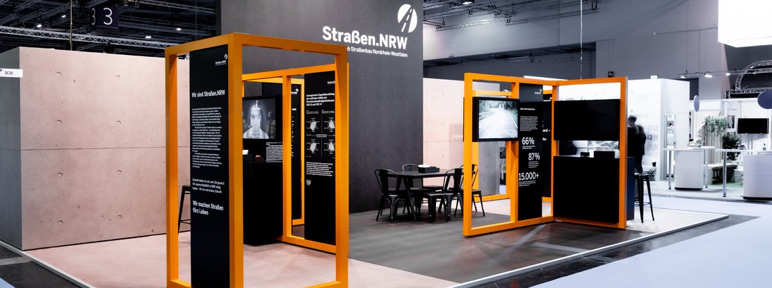 VISUELL Scenography: Straßen.NRW stand at InfraTech 2026: A view of the stand; the striking orange frame structure divides the 50-square-metre stand area into various themed zones featuring integrated screens and information panels.