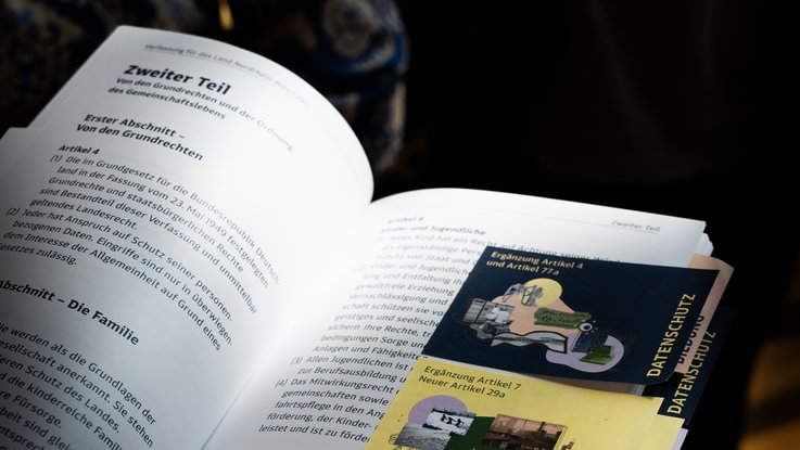 VISUELL Szenografie: Wanderausstellung „Jubiläum. 75 Jahre Verfassung NRW“ für die Stiftung Haus der Geschichte Nordrhein-Westfalen VISUELL Szenografie: Wanderausstellung: Geöffnetes Exemplar der NRW-Verfassung mit eingelegten Themenkarten zu Datenschutz, Bildung und Grundrechten.
