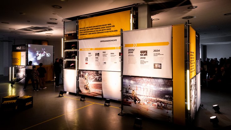 VISUELL Szenografie: Ausstellung: Überblick über mehrere Ausstellungswürfel mit großflächigen Texttafeln und Fotos; Besucherinnen und Besucher betrachten die Inhalte, während Projektionen historische Szenen ergänzen.