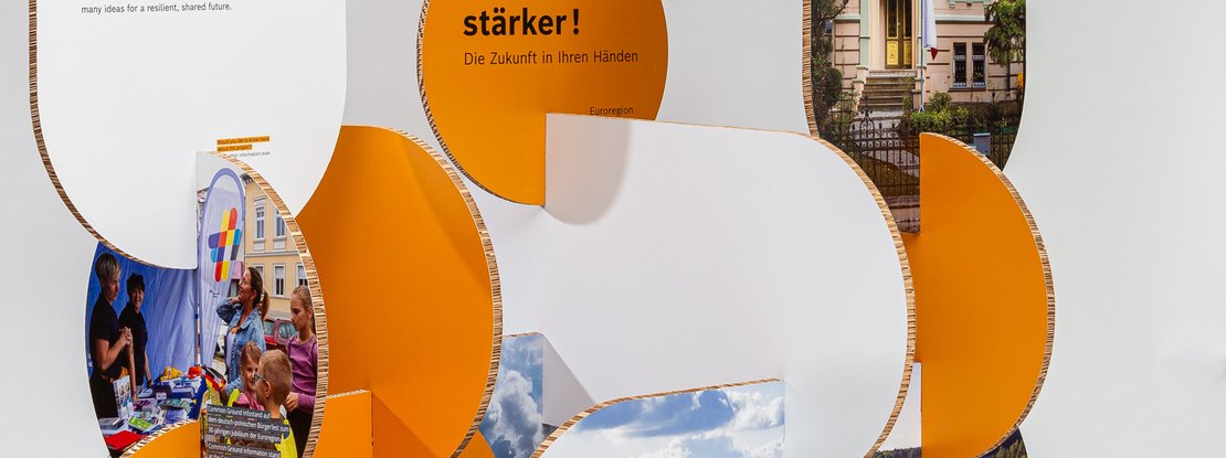 VISUELL Scenography: Robert Bosch Stiftung GmbH travelling exhibition: a close-up view of an organically shaped exhibition stand made of orange and white honeycomb panels; integrated display panels show scenes from events and landscapes, with lettering above that ends with “…stronger! The future is in your hands”.