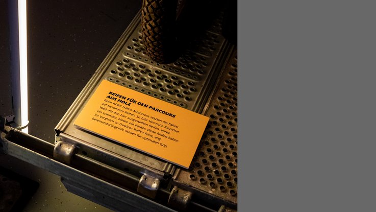 VISUELL Szenografie: Ausstellung: Detail einer gelben Texttafel zu speziellen Reifen für den Parcours; darüber die Exponate mit grobstolligen Rädern, eingerahmt von Lichtakzenten.