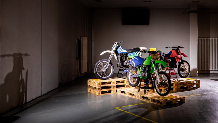 VISUELL Szenografie: Ausstellung: Drei Motorräder unterschiedlicher Baujahre auf gestapelten Paletten, einzeln angestrahlt; ihre Schatten zeichnen sich groß auf der Wand ab.
