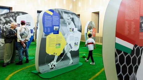 VISUELL Szenografie: EURO Legends Stuttgart: Eine Frau und ein Mann stehen zwischen überlebensgroßen Aufstellern von Fußballspielern, ein Junge dribbelt mit einem Ball durch den Parcours.