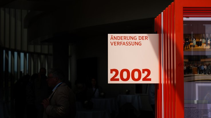 VISUELL Szenografie: Wanderausstellung „Jubiläum. 75 Jahre Verfassung NRW“ für die Stiftung Haus der Geschichte Nordrhein-Westfalen VISUELL Szenografie: Wanderausstellung: Detail eines Ausstellungselements mit der Aufschrift „Änderung der Verfassung 2002“. Rotes Rahmenelement im Vordergrund, Personen im Hintergrund im Halbdunkel.