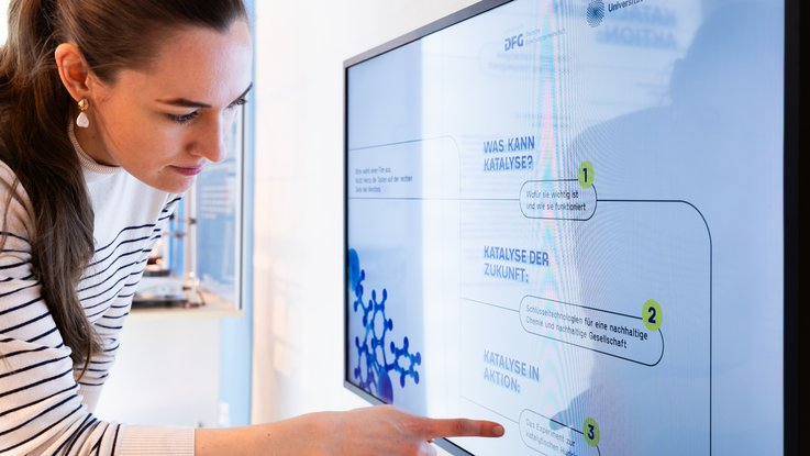 VISUELL Szenografie: Wissenschaftsausstellung: Nahaufnahme einer Besucherin die vor einem Bildschirm steht und auf Elemente der dort abgespielten Informationsgrafik zeigt.