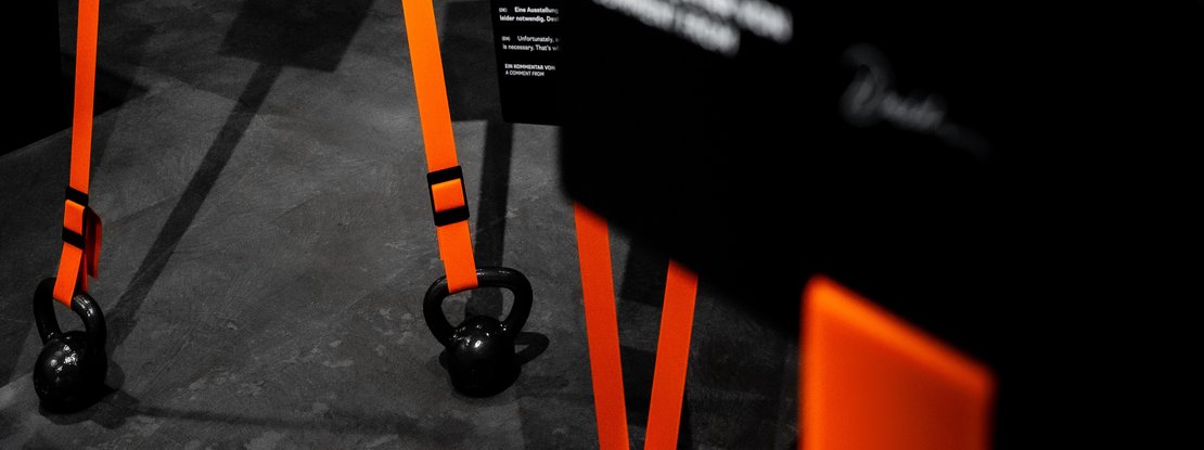 VISUELL Szenografie: Sonderausstellung: Anti Anti 2.0: Vertikale Installation mit leuchtend orangefarbenen Bändern und Gewichten (Kettlebells); daran befestigte Informationstafeln markieren eine Station innerhalb der Dauerausstellung und setzen visuelle Impulse im Raum. 