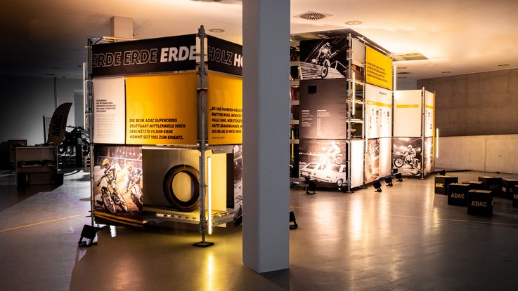 VISUELL Szenografie: Ausstellung: Modulare Kuben mit Bannern, Fotos und Texten formen eine offene Inszenierung; markante Schwarz-Gelb-Gestaltung prägt das Gesamtbild.