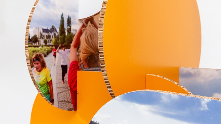 VISUELL Szenografie: Wanderausstellung „Common Grounds – Über Grenzen mitgestalten“ für die Robert Bosch Stiftung GmbH VISUELL Szenografie: Wanderausstellung Robert Bosch Stiftung GmbH: Detailansicht zusammengesteckter Ausstellungselemente mit großformatigen Bildmotiven von Menschen und Landschaft; orangefarbene Trägerformen schaffen Tiefe und Rhythmus.