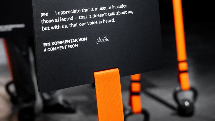 VISUELL Szenografie: Sonderausstellung: Anti Anti 2.0: Nahaufnahme eines Textträgers mit orangefarbenem Band im Hintergrund; Kommentare erscheinen unmittelbar am Objekt.