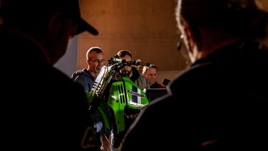 VISUELL Szenografie: Ausstellung: Besuchergruppe betrachtet aus der Nähe ein historisches grünes Motorrad; die konzentrierten Blicke lassen die Faszination für Technik spürbar werden.