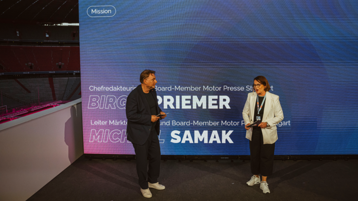VISUELL Szenografie: Event „NEXT LEVEL“: Zwei Personen stehen auf der Bühne vor einer großen LED-Leinwand in der Allianz Arena, die Leinwand zeigt den Titel „Mission“.