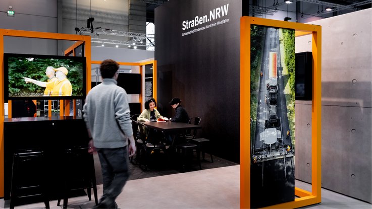 VISUELL Scenography: Straßen.NRW stand at InfraTech 2026: People are seated at a table in the stand’s dialogue area, surrounded by a carefully curated combination of photographs, graphics and moving images.