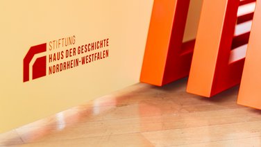 VISUELL Szenografie: Wanderausstellung „Jubiläum. 75 Jahre Verfassung NRW“ für die Stiftung Haus der Geschichte Nordrhein-Westfalen VISUELL Szenografie: Wanderausstellung: Detail der orangefarbenen Lamellenstruktur mit dem roten Logo der Stiftung Haus der Geschichte Nordrhein-Westfalen auf gelber Fläche.