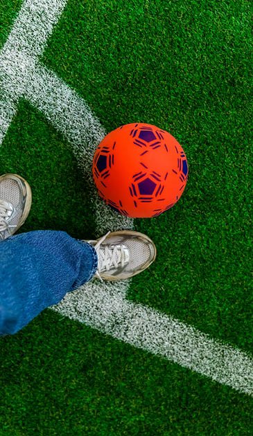 VISUELL Szenografie: EURO Legends Stuttgart: Ein roter Fußball liegt auf einem grünen Fußballfeld aus Kunstrasen mit weißen Markierungen, daneben sieht man die Turnschuhe einer Person.