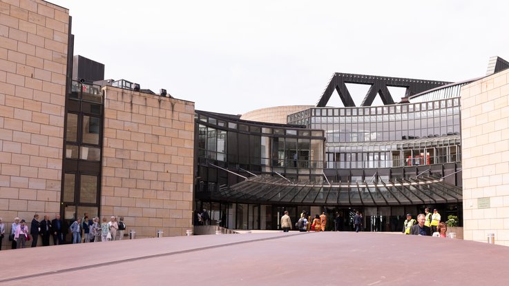 VISUELL Szenografie: Wanderausstellung „Jubiläum. 75 Jahre Verfassung NRW“ für die Stiftung Haus der Geschichte Nordrhein-Westfalen VISUELL Szenografie: Wanderausstellung: Eingangssituation des Landtags NRW, Gruppen von Besucher:innen stehen und gehen über den Vorplatz, im Hintergrund die gläserne Fassade des Gebäudes.