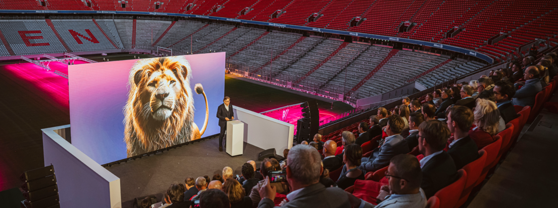 VISUELL Szenografie: Event „NEXT LEVEL“: Die Zuschauenden sitzen auf der Tribüne, eine Person steht hinter einem Rednerpult vor der großen LED-Leinwand. Im Hintergrund sieht man die leeren ränge der Allianz Arena und das Spielfeld.