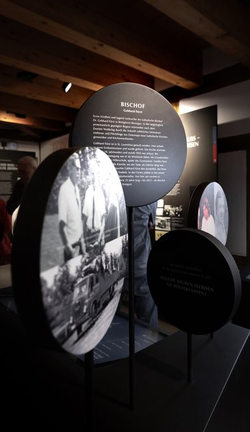 VISUELL Scenography: Permanent exhibition at Hornmoldhaus: Free-standing round picture and text panels on slender steles with portraits and biographical information, including a panel on Bishop Gebhard Fürst. The dark design contrasts with the historic wooden beam ceiling of the room.
