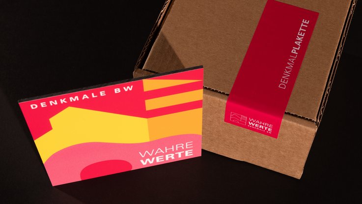 VISUELL Kommunikationsdesign: Draufsicht auf die Denkmalplakette des Landesamts für Denkmalpflege, welche am dazugehörigen Karton mit abgestimmtem Verpackungssiegel lehnt