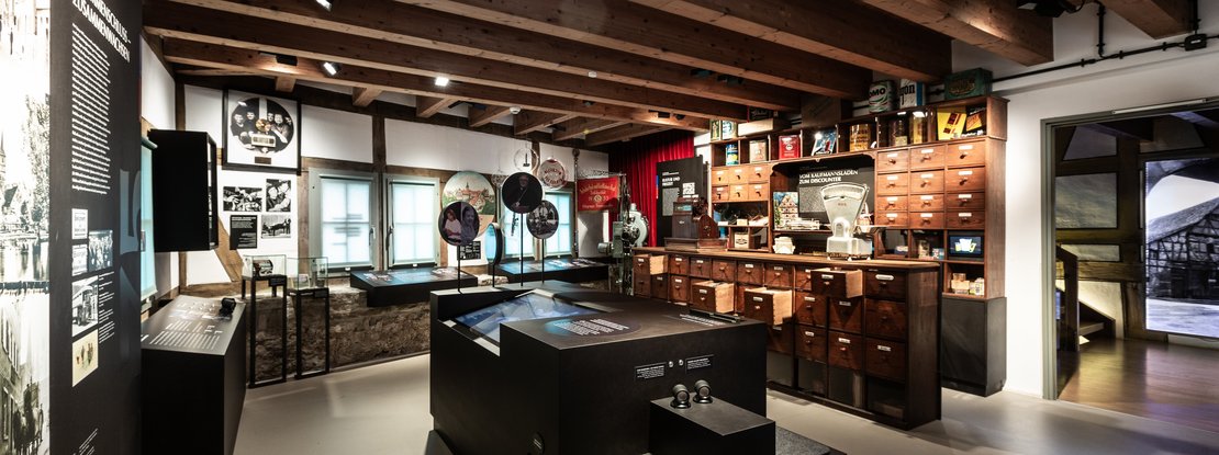 VISUELL Scenography: Permanent exhibition Hornmoldhaus: Historical exhibition space with exposed wooden beam ceiling, central black media table and surrounding text and image panels. Integrated historical merchant's cabinet on the right with numerous drawers and shop scales, supplemented by exhibits and round picture panels on the history of the town.