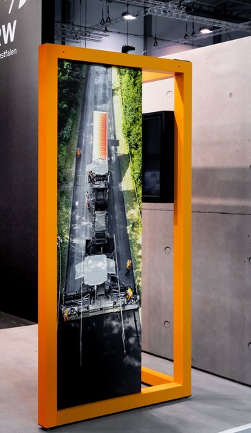 VISUELL Scenography: Straßen.NRW stand at InfraTech 2026: a graphic display in an orange frame shows a bird’s-eye view photograph depicting roadworks and road maintenance vehicles