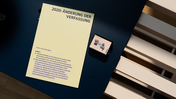 VISUELL Szenografie: Wanderausstellung „Jubiläum. 75 Jahre Verfassung NRW“ für die Stiftung Haus der Geschichte Nordrhein-Westfalen VISUELL Szenografie: Wanderausstellung: Detailaufnahme der Tafel „2020: Änderung der Verfassung“. Text zu Artikel 1 und 3 der NRW-Verfassung auf gelbem Hintergrund mit kleiner grafischer Illustration.