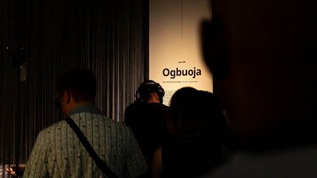 VISUELL Szenografie: Intervention Afrika: Viele Menschen stehen vor einem Banner mit dem Titel der Intervention von Sett Ofili „Ogbuoja: The Story of a Flute Player“