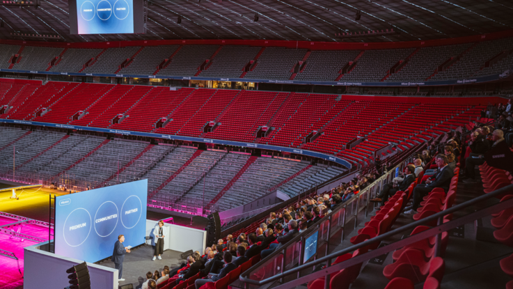 VISUELL Szenografie: Event „NEXT LEVEL“: Die Zuschauenden sitzen vor der Leinwand auf der Tribüne, zwei Personen stehen vor der großen LED-Leinwand. Im Hintergrund sieht man die leeren ränge der Allianz Arena und das Spielfeld.